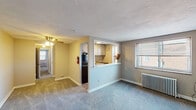 This single-floor residence offers a practical and well-organized layout across a comfortable footprint. The main living area is bright and open, finished with plush carpeting in a neutral tone and painted in a soft gray palette. A ceiling fan provides overhead lighting and airflow, while a classic white radiator positioned beneath a window ensures consistent heating. Natural light filters in through blinds-fitted windows, offering views of the surrounding neighborhood.

The kitchen is thoughtfully integrated via a pass-through opening, creating a connected feel between the cooking and living spaces. White cabinetry lines the kitchen walls, and a dark refrigerator anchors the space alongside a countertop range. This semi-open design enhances the sense of flow throughout the home.

A hallway connects the living area to the rest of the unit, leading to two carpeted bedrooms. Each bedroom features neutral-toned walls, a window with blinds that admits natural light, and its own white radiator. The rooms offer a calm, understated atmosphere well-suited for rest or use as a home office.

The bathroom is compact and functional, featuring a tub-shower combination with a decorative curtain, a toilet, and wood-look flooring that adds a warm contrast to the otherwise neutral finishes throughout the unit.
