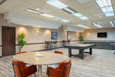 Located at 6617 Odana Rd in Madison, Wisconsin, this generously sized single-floor amenity space offers a well-appointed, open-plan layout designed for social interaction and leisure. The interior is brightened by a drop-tile ceiling with large skylight-style translucent panels, which flood the space with diffused ambient light, complemented by recessed fixtures and warm wall sconces throughout.

The lounge area serves as a central focal point, anchored by a substantial sectional sofa in slate gray with an oversized tufted ottoman, paired with a set of curved, emerald-green velvet accent chairs. Colorful local artwork and decorative prints add personality to the neutral-toned walls, which feature a distinctive horizontal accent stripe.

Adjacent to the lounge, a round dining table surrounded by rust-orange upholstered chairs provides a casual gathering spot. Further into the space, a pool table and foosball table define a dedicated game zone, accompanied by bar-height seating with russet-toned stools for spectators.

A built-in shelving unit and a wall-mounted flat-screen television with a sleek console table create a media nook near one end of the room. Light wood-look flooring runs throughout, contrasting warmly with the patterned area rugs. The overall ambiance is modern, inviting, and purposefully designed for community use.
