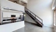 This two-floor urban loft masterfully combines industrial heritage with contemporary design. The main level greets you with polished concrete floors, soaring ceilings, and exposed silver HVAC ductwork that runs throughout, reinforcing the raw, warehouse-inspired aesthetic. The kitchen is well-appointed with white shaker cabinetry, dark granite countertops, stainless steel appliances, and a kitchen island — all set against a backdrop of original exposed brick that adds warmth and texture to the space. A bold steel-and-wood open staircase connects the two levels, serving as both a functional and architectural centerpiece. The lower level also includes a full bathroom with polished concrete flooring, a glass-enclosed shower, and a floating vanity. Ascending to the upper level, rich hardwood flooring transitions the aesthetic toward a warmer, more residential feel. The spacious upper room features track lighting, a ceiling fan, and an open view overlooking the dramatic double-height brick wall below, framed by large windows that flood the space with natural light. The upper level is further complemented by a generously sized bathroom with a dual-sink vanity, a walk-in closet with built-in shelving, and an in-unit laundry room equipped with a full-size washer and dryer.