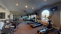 This fitness center is a comprehensively equipped athletic space designed to support a wide range of workout routines. The room features vaulted ceilings that lend an open, airy feel, while a ceiling fan provides additional ventilation during workouts. Recessed lighting supplements the abundant natural light streaming through multiple large windows, including a striking arched window that frames views of the surrounding outdoor greenery.

The cardio zone is well-stocked with multiple treadmills, elliptical trainers, and stationary and recumbent bikes, all positioned to take advantage of the natural light and outdoor views. A wall-mounted television keeps users entertained during extended cardio sessions. A full-length mirror spans one wall, enhancing the sense of space while allowing users to monitor their form.

The strength training area includes a multi-function cable and weight machine, an adjustable workout bench, and a varied collection of kettlebells in multiple weights, neatly arranged along the floor. Additional free weights and a weight rack round out the strength equipment offerings. The flooring combines durable carpet in the main exercise areas with hardwood-style flooring in the strength section, providing both comfort and functionality throughout the space.