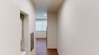 This single-floor unit offers a clean, contemporary design with a cohesive aesthetic carried throughout each space. Warm wood-tone laminate flooring runs continuously across the unit, complementing the neutral, light-toned walls and white trim that give the interior a bright, airy feel.

The kitchen is a standout feature, equipped with a full suite of stainless steel appliances, including a refrigerator, electric range, over-the-range microwave, and dishwasher. Light wood flat-front cabinetry extends to the ceiling, providing generous storage, while white countertops offer a clean, modern contrast. A single-basin sink anchors the workspace, making the kitchen both functional and visually cohesive.

A connecting hallway leads from the entry area, where a built-in panel and utility access are visible, into the main living space. The bedroom features a sliding-door closet with wire shelving, recessed lighting, and a window fitted with a roller shade that allows for natural light while maintaining privacy. The open layout of the main room provides flexible use of space, with recessed ceiling lights ensuring consistent illumination throughout.

Overall, the unit presents a modern, move-in-ready interior with practical finishes, efficient use of space, and a streamlined design that balances functionality with a clean, contemporary aesthetic.