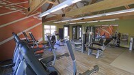 This fitness center offers a comprehensive workout environment designed to accommodate a variety of exercise routines. The space is defined by its striking architectural character, featuring exposed wooden roof trusses and vaulted ceilings that lend an open, airy feel. Bold orange and olive green accent walls add visual energy to the room, complementing the dynamic atmosphere of the facility.

The flooring consists of wood-toned planks throughout, providing a durable and clean surface suited for heavy equipment. Natural light enters through windows along the perimeter, brightening the space alongside overhead fluorescent lighting mounted between the exposed beams.

The gym is outfitted with a solid selection of cardio equipment, including treadmills, ellipticals, and stationary bikes positioned toward the front and sides of the room. Strength training equipment, including cable machines and weight benches, occupies the remaining floor space. A large wall mirror on one side enhances the sense of depth while allowing users to monitor their form. The overall layout is practical and well-organized, making efficient use of the available floor space for a functional, full-body training experience.