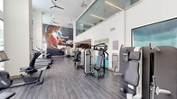 Located at 480 N Orange Ave in the heart of downtown Orlando, this fitness center at Central Station on Orange is a well-appointed, single-floor facility offering a generous workout environment. The space is defined by soaring double-height ceilings, expansive floor-to-ceiling windows that flood the interior with natural light, and views of the surrounding urban landscape and pool area.

The flooring throughout consists of durable wood-look planks, lending a clean and modern aesthetic that complements the bright white walls. A striking large-scale mural of yoga practitioners serves as a bold focal point, reinforcing the wellness-focused atmosphere of the space.

The cardio zone features a row of commercial-grade treadmills and other aerobic machines aligned along the windowed wall, maximizing natural light during workouts. The strength training area is well-equipped with a comprehensive selection of cable machines, resistance equipment, and adjustable benches, accommodating a wide range of exercise routines.

Additional highlights include a mirrored free weights section with wall-mounted TVs, a teal open-shelving unit for storage near the entrance, and a contemporary open-riser staircase with metal railings. Ceiling fans throughout help maintain airflow, and an upper-level mezzanine overlooks the main floor, adding architectural depth to the overall design.