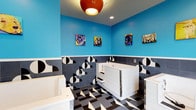This cheerful and purpose-built pet wash facility is designed with both function and personality in mind. The space features multiple white, walk-in style dog bathing tubs equipped with handheld sprayer attachments and built-in steps, making them accessible for pets of varying sizes. Each tub is fitted with chrome fixtures and thoughtfully positioned for easy handling.

The interior design is bold and eye-catching. The lower walls are clad in dark gray tiles adorned with a striking black-and-white geometric circle pattern, while the upper walls are painted in a vivid sky blue that energizes the entire room. A white dividing border cleanly separates the two zones, adding a crisp visual contrast.

The floor continues the playful theme with a dynamic black, white, and gray tile arrangement in an abstract geometric pattern. Overhead, a retro-inspired orange pendant light fixture anchors the space alongside recessed ceiling lights, ensuring bright, even illumination throughout.

Adding warmth and character to the walls, a curated collection of colorful, pop-art style painted portraits of various dog breeds is displayed, reinforcing the pet-friendly spirit of the space. A set of glass-paned double doors at one end floods the room with natural light and provides an exterior view, enhancing the open, welcoming atmosphere of this well-appointed grooming facility.
