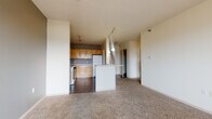 This single-floor residence is designed with an open-concept layout that seamlessly connects the kitchen and living area. The kitchen is well-equipped, featuring warm honey-toned wood cabinetry extending to the upper walls for ample storage, dark gray accent walls, dark hardwood-style flooring, and white appliances including an electric range, refrigerator, and dishwasher. A peninsula with a white countertop extends into the living space, providing a natural transition between the two areas and offering additional seating or workspace. Track lighting illuminates both the kitchen and the adjoining living area.

The living area is generously sized, featuring neutral-toned carpet and light-colored walls that create a bright, open atmosphere. A taupe accent wall adds a subtle design element, and the space accommodates a variety of furniture arrangements.

The bedroom is a comfortable size with soft carpet flooring, neutral walls, and a window that allows natural light to fill the room. The bathroom includes a wide vanity with light wood cabinetry, a white countertop, a large mirror, and dark hardwood-style flooring, offering a clean and functional design.