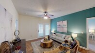 This single-floor residence offers a thoughtfully designed layout that balances comfort and style across its key living spaces. The living room serves as the heart of the home, featuring rich hardwood-style flooring, a textured ceiling with a ceiling fan, and a striking teal accent wall adorned with abstract artwork. A set of glass-paned French doors floods the space with natural light and provides outdoor access, creating an open, airy feel. The adjacent dining area is anchored by an elegant chandelier and continues the warm hardwood flooring, offering an intimate setting for meals. The kitchen is well-equipped with warm-toned cabinetry, light granite-style countertops, stainless steel appliances, and a double-basin sink — all set over durable hardwood-style flooring. The primary bedroom features plush carpeting, a bold dark teal accent wall, ceiling fan, and ample natural light from large windows with white curtains. A second bedroom mirrors this comfort with soft carpet, a ceiling fan, and its own accent wall in a calming blue hue. Two bathrooms complete the layout, each featuring granite countertops, white cabinetry, and hardwood-style flooring, with one offering a large vanity mirror with Hollywood-style lighting.