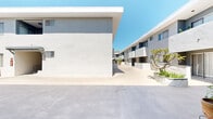 Located at 111 N Eucalyptus Ave in Inglewood, California, this expansive open-air courtyard spans a substantial ground-level footprint, configured in an L-shaped layout as visible from the aerial scan. The complex is defined by its clean, white stucco exterior walls and a modern, minimalist architectural style that reflects Southern California's characteristic aesthetic.

The courtyard runs as a wide, open concrete corridor flanked on both sides by two-story residential building wings, creating a sense of enclosure while remaining fully open to the sky. Natural light floods the space throughout the day, bathing the smooth concrete walkways in warm Southern California sunshine. Potted trees and planter beds featuring tropical and ornamental plants are positioned throughout the courtyard, adding touches of greenery and visual interest to the otherwise neutral palette.

Stairways with metal railings connect the ground level to the upper floor walkways, and covered pergola-style overhangs provide shaded entry points at various building access areas. Unit entry doors are evenly spaced along the courtyard-facing walls, with sliding windows and vertical blinds consistent across the facade. Security gates and fire safety equipment are also present at key access points. The overall ambiance is bright, orderly, and open, characteristic of a well-maintained multi-unit residential courtyard property.