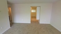 This single-floor space presents a clean, functional layout designed with simplicity and practicality in mind. The main living area is finished with wall-to-wall neutral-toned carpeting that adds warmth underfoot, while the crisp white walls create a bright and open atmosphere throughout. A wide doorway connects the carpeted living space to an adjoining room finished with light-colored ceramic tile flooring, offering a clear distinction between living and utility zones. The tiled room benefits from its own natural light source via a window fitted with blinds, adding to the overall brightness of the interior. The transition between carpet and tile is clean and well-defined, reflecting a straightforward but thoughtful design approach. Electrical outlets are conveniently positioned along the walls of the main area, ensuring practical usability throughout the space. The overall aesthetic is neutral and move-in ready, with white walls and light flooring tones that provide a versatile backdrop suitable for a variety of interior styles. The single-story layout ensures ease of access and navigation throughout, making this a highly functional and accessible space.