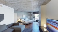 This thoughtfully designed single-floor space combines industrial and contemporary aesthetics to create a refined and comfortable environment. The most striking architectural feature is the exposed raw concrete ceiling, which lends an urban, loft-like character to the room while contrasting beautifully with the warm tones of the herringbone-patterned hardwood flooring beneath.

The sleeping area is anchored by a generously sized bed dressed in crisp white linens, flanked by bedside tables with warm ambient lighting. Adjacent to the bed, a cozy lounge area offers a plush gray sofa and ottoman, arranged over a soft area rug, creating a relaxed and inviting retreat within the space.

A sliding glass door at the far end of the room floods the interior with natural light and provides direct access to an outdoor area, where a small seating arrangement takes advantage of the exterior views. The transitional zone near the glass door features additional seating, perfect for quiet relaxation.

Cool gray walls complement the industrial ceiling and work harmoniously with the warm wood floors and soft furnishings. A wall-mounted air conditioning unit and thoughtfully placed modern light fixtures complete the space. A partial yellow-accented wall and media console add subtle pops of color, enhancing the room's contemporary character.