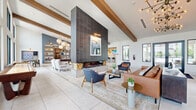 Located at 2600 Lake Betty Blvd in Apopka, Florida, this generously scaled single-floor community clubhouse delivers a sophisticated, resort-inspired atmosphere across a spacious open layout. The centerpiece of the main lounge is a striking floor-to-ceiling dark tile fireplace flanked by curated seating arrangements, including leather sofas and mid-century accent chairs, all anchored by large-format light stone tile flooring that flows throughout the entire space. Soaring vaulted ceilings with exposed wood beams and an array of contemporary pendant and chandelier lighting add warmth and architectural character to the open interior. Large black-framed windows flood the space with abundant natural light while offering views of the surrounding Florida landscape. A dedicated recreation area houses a full-size billiards table alongside additional lounge seating, while a built-in bookshelf with accent lighting provides a relaxed reading nook. The kitchen and social bar area features sleek sage cabinetry, a dramatic marble-style countertop island with seating, a wood-paneled ceiling detail, and pendant lighting with brass accents. A live-edge communal dining table nearby adds an organic, natural element. Glass-walled private offices and meeting rooms provide functional workspace, while a cozy game nook with a wall-mounted board game and natural light rounds out this thoughtfully designed community gathering space.