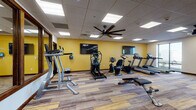 This fitness center offers a comprehensive workout facility spread across a single floor, thoughtfully designed to accommodate a wide range of exercise needs. The main workout area features warm yellow accent walls, multi-tone wood-look flooring, and a drop-panel ceiling outfitted with recessed panel lighting and a large ceiling fan for optimal air circulation. Two wall-mounted flat-screen TVs keep exercisers entertained throughout their sessions.

The cardio zone is well-stocked with multiple treadmills, an elliptical trainer, a stair climber, and stationary bikes, including both upright and recumbent options. A large window along one wall floods the treadmill area with natural light and provides an outdoor view. The strength training section includes a multi-station cable machine, a full rack of dumbbells in graduated weights, and a weight bench, with floor-to-ceiling mirrors amplifying the sense of space.

A separate studio room provides a quieter, open-plan area ideal for cycling classes or group exercise, equipped with spin bikes and a wall-mounted rack of medicine balls and exercise mats. The light gray flooring and neutral walls give this room a clean, versatile atmosphere. Together, these spaces create a fully functional and inviting fitness environment suitable for users of all fitness levels.