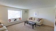 This single-floor space features a living room that serves as its central gathering area. The room is finished with wall-to-wall carpeting in a neutral tone, contributing to a warm and comfortable atmosphere. Smooth, light-colored walls complement the textured ceiling overhead.

A large window along one wall allows abundant natural light to fill the room, offering views of the surrounding outdoor environment. A wall-mounted air conditioning unit is positioned above the window, while baseboard heating runs along the lower perimeter of the walls, ensuring year-round climate control.

The living area comfortably accommodates a cushioned sofa accented with patterned throw pillows and a dark-framed rectangular coffee table, demonstrating its functional layout. A framed artwork piece on the wall adds a subtle decorative touch. The windowsill provides a small display ledge, currently hosting a few decorative objects.

Overall, the space presents a clean, understated aesthetic with a practical and livable design, well-suited for everyday use.