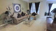 This single-storey space showcases a thoughtfully designed living room that balances comfort and sophistication. The sitting area is anchored by a generously sized taupe sofa adorned with an arrangement of dark chocolate and neutral accent cushions, paired with a coordinating striped armchair featuring a bold diamond-patterned cushion. A sleek glass-and-chrome coffee table serves as the centrepiece, offering both functionality and visual lightness to the room.

The flooring is finished in plush neutral carpet, contributing to the warm and inviting ambiance. Walls are painted in a soft sage-green tone, providing a calm backdrop that complements the dark accents throughout. Crown moulding runs along the ceiling line, adding an architectural detail that enhances the room's refined character.

Natural light enters through a large window fitted with white plantation shutters, while floor-length black drapes frame the window dramatically, creating a striking contrast. A decorative wall sconce and a table lamp provide layered ambient lighting. A large framed abstract artwork on the wall adds a contemporary artistic touch, completing the cohesive and polished aesthetic of the space.