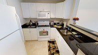 This single-floor residence offers a thoughtfully designed living space with a classic and functional aesthetic throughout. The kitchen is a standout feature, showcasing crisp white shaker-style cabinetry that extends to the upper walls, providing generous storage and a bright, airy feel. Dark granite countertops offer a striking contrast against the white cabinets, adding a touch of elegance to the space. The kitchen is equipped with white appliances, including an electric range with oven, an over-the-range microwave, and a stainless steel double-basin sink, all seamlessly integrated into the layout. The U-shaped kitchen design maximizes counter space and workflow efficiency, making it practical for everyday cooking and entertaining. Light tile flooring ties the space together, complementing the overall clean and cohesive color palette. A decorative area rug adds warmth and a personal touch to the kitchen floor. The cabinetry hardware and thoughtful use of counter space reflect an attention to detail that carries throughout the residence. Overall, the space presents a well-maintained, move-in-ready environment with a timeless design that balances style and practicality.