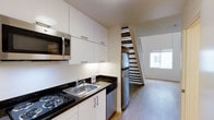 This two-floor space is designed with functionality and clean aesthetics in mind. The kitchen is efficiently laid out, featuring dark granite countertops that contrast sharply with white cabinetry extending to the ceiling, providing generous storage. Stainless steel appliances, including a full-size refrigerator, built-in microwave, and a coil-burner stove, equip the kitchen for everyday use. A stainless steel sink with a modern faucet is integrated into the counter, alongside a dishwasher tucked neatly beneath. Light wood flooring extends through the space, adding warmth to the otherwise crisp, neutral palette of white walls and cabinetry. Adjacent to the kitchen, an open staircase with a distinctive alternating-tread design connects the two floors, serving as both a functional and architectural feature. A window near the staircase allows natural light to filter into the living area, brightening the open lower level. The overall ambiance is clean, compact, and modern, making efficient use of vertical space across both floors.