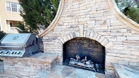 This outdoor amenity space is thoughtfully designed to offer a comprehensive al fresco living experience. At its centerpiece is a generously sized, freeform swimming pool with a smooth concrete surround, lined with multiple lounge chairs positioned to capture ample sunlight throughout the day. A covered pavilion with a pergola structure provides a shaded retreat at the pool's edge.

The focal point of the outdoor entertaining area is a striking stacked-stone fireplace, constructed with warm-toned natural stone in a classic arched design. The chimney rises with a decoratively curved profile, and the hearth is set atop a flagstone patio that adds a rugged, natural texture underfoot. Flanking seating ledges integrate seamlessly into the stonework, creating a welcoming gathering spot.

Adjacent to the fireplace is a built-in outdoor kitchen station, featuring a stainless steel gas grill mounted within a matching stacked-stone surround. Stone planter walls with seasonal landscaping define the outdoor kitchen area, adding color and natural separation between zones. Decorative street-style lighting enhances usability into the evening hours, while mature trees and manicured shrubs provide greenery and a sense of privacy throughout the space.