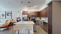 This single-floor residence showcases a contemporary design with clean lines, wood-look flooring, and a neutral color palette throughout. The open-concept kitchen serves as a central highlight, featuring walnut-toned upper cabinetry, light gray lower cabinets, and a full suite of stainless steel appliances including a French door refrigerator, range, microwave, and dishwasher. A large white-countertop island with bar seating provides additional prep space and casual dining. Pendant lighting and a track light fixture illuminate the kitchen effectively.

Adjacent to the kitchen, the dining area accommodates a glass-top table with chrome-framed chairs beneath bold abstract artwork. The living room receives natural light through roller-shaded windows and offers a comfortable, well-proportioned lounge space.

Both bedrooms are quietly appointed with large windows fitted with roller shades, wood-look flooring, and wall art. The bathroom features a wide undermount sink with a modern faucet, a large vanity mirror, and a glass-enclosed walk-in shower. Grab bars are present, indicating accessibility-conscious design. Throughout, the space maintains a cohesive modern aesthetic with thoughtful detailing.