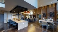 Located on Foster Street in Durham, NC, this single-floor space delivers a striking industrial-modern aesthetic that blends raw materials with refined design. The open-plan layout is anchored by soaring ceilings that amplify the sense of volume and grandeur throughout. Exposed brick walls provide a warm, textural contrast against sleek dark surfaces, while polished concrete floors run seamlessly across the entire space, reinforcing its contemporary character.

The centerpiece of the space is a generously sized gourmet kitchen area featuring multiple islands — one finished in light granite and another in dark stone — offering ample surface area for food preparation and casual seating. Industrial-style pendant lighting, including bold matte black fixtures and delicate globe pendants suspended at varying heights, creates dramatic visual layering and warm, inviting illumination. Full-sized stainless steel appliances, custom cabinetry, and a dedicated bar seating area make this kitchen as functional as it is stylish.

Curated artwork adorns the walls, adding cultural depth and personality to the environment. The overall ambiance is bold yet welcoming, combining urban industrial elements with thoughtful design details that make this a versatile and memorable communal destination.