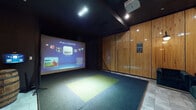 Located at 1333 N Santa Fe Ave in Vista, California, this single-floor indoor golf simulator venue offers a refined and immersive entertainment experience. The space is defined by rich, floor-to-ceiling wood-paneled walls that lend a warm, upscale ambiance throughout. The primary feature is a dedicated golf simulator bay equipped with a Full Swing Golf system, one of the most advanced simulator platforms available, capable of recreating the world's most iconic courses with high-fidelity graphics. The hitting area is outfitted with a multi-toned turf mat and is illuminated by focused overhead lighting, creating an atmosphere reminiscent of a private golf suite. A large projection screen dominates the far wall of the simulator bay, providing an immersive visual experience for players. Adjacent to the simulator, the space transitions into a lounge and bar area, where a decorative wooden barrel and a sleek black metal bar cart stocked with glassware add a sophisticated, speakeasy-inspired touch. The lounge area features plush green velvet tufted seating and framed wall art, enhancing the upscale aesthetic. Tile flooring anchors the non-turf areas, while recessed and accent lighting throughout creates a moody, intimate atmosphere ideal for private events or recreational play.