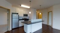 This single-floor space showcases a well-designed open-concept kitchen and living area. The kitchen is equipped with stainless steel appliances, including a full-size refrigerator, a gas range with overhead microwave, and ample white cabinetry providing generous storage. Dark countertops contrast cleanly against the white cabinetry and walls, creating a modern, polished aesthetic. A central island with matching dark countertops serves as a functional workspace and gathering point, illuminated by two stylish pendant lights that hang at varying heights, adding visual interest to the space. The kitchen benefits from both recessed and overhead lighting, ensuring the area is well-lit. Dark hardwood flooring runs throughout, grounding the space with warmth and continuity. A hallway visible in the background hints at additional rooms beyond the main living area, with warm-toned lighting offering a welcoming transition. The overall layout is practical and contemporary, balancing functionality with clean, minimalist design elements that make the space feel open and well-organized.