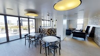 This contemporary single-floor communal space is thoughtfully designed to encourage social interaction and relaxation. The main lounge area features round café-style tables paired with sleek black chairs, arranged to maximize the open, airy feel of the room. Striking oversized circular pendant lights in gold and black hang from the smooth white ceiling, serving as bold focal points that add warmth and visual interest to the space.

Large windows line the walls, flooding the interior with natural light and offering views of an outdoor terrace or pergola area, creating a seamless connection between indoor and outdoor environments. The flooring throughout is a neutral large-format tile, lending a clean and modern aesthetic.

A bar or counter area anchors the back of the room, featuring colorful mosaic-tiled bar fronts and accompanied by bar-height seating, making it an ideal spot for casual gatherings. Additional pendant lighting above the bar provides focused illumination in this area.

To one side, a lounge seating zone with dark upholstered sofas and chairs offers a more relaxed setting, separated subtly by architectural columns that define the space without enclosing it. Together, these zones create a versatile, welcoming environment suited for both casual socializing and informal meetings.