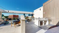 This stylish rooftop terrace offers an exceptional open-air retreat with a well-appointed layout designed for both relaxation and entertaining. The space is finished with large-format porcelain tile flooring throughout, lending a clean and contemporary aesthetic that complements the crisp white architectural exterior panels and textured concrete accent walls.

A standout outdoor kitchen anchors one side of the terrace, featuring a sleek marble-topped counter with gold veining, an integrated sink, and under-counter refrigeration — ideal for alfresco dining and hosting. Nearby, a built-in linear fireplace is set into a smooth stucco wall, creating a warm focal point flanked by casual seating and a small bistro table.

Distinct zones throughout the terrace accommodate a variety of uses: a lounge area with woven accent chairs and colorful furnishings invites casual conversation, while a dedicated dining area with a modern table and contemporary chairs is perfect for group gatherings. String lights overhead add ambient evening appeal.

Glass-panel railings along the perimeter ensure unobstructed panoramic views of the surrounding urban skyline, with city buildings stretching toward the horizon beneath bright blue skies. Lush planters add touches of greenery, softening the modern hardscape. The terrace is accessed via a dedicated stairwell entry, completing this versatile and visually striking outdoor amenity.