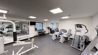 This fitness center offers a well-organized and fully equipped workout environment designed to support a range of exercise routines. The space is finished with a durable, speckled rubber flooring that provides both safety and comfort underfoot, while the crisp white walls and dark ceiling create a sleek, modern aesthetic. Bright recessed panel lighting illuminates the entire room evenly, ensuring excellent visibility throughout all areas of the facility. A large wall-mounted mirror along one side enhances the sense of space and allows users to monitor their form during workouts. The cardio section is well-stocked with upright and recumbent exercise bikes, along with an elliptical trainer, catering to a variety of cardiovascular fitness preferences. A dedicated free weights area features a tiered dumbbell rack with a comprehensive range of weights, accompanied by a flat workout bench for strength training exercises. Additional equipment, including a cable machine and a stationary bike with a monitor, rounds out the strength and conditioning offerings. Two windows allow natural light to filter into the space, complementing the artificial lighting and contributing to an open, energizing atmosphere. Overall, the fitness center presents a clean, functional, and inviting environment suitable for a broad spectrum of fitness activities.