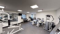 This fitness center offers a well-organized and fully equipped workout environment designed to support a range of exercise routines. The space is finished with a durable, speckled rubber flooring that provides both safety and comfort underfoot, while the crisp white walls and dark ceiling create a sleek, modern aesthetic. Bright recessed panel lighting illuminates the entire room evenly, ensuring excellent visibility throughout all areas of the facility. A large wall-mounted mirror along one side enhances the sense of space and allows users to monitor their form during workouts. The cardio section is well-stocked with upright and recumbent exercise bikes, along with an elliptical trainer, catering to a variety of cardiovascular fitness preferences. A dedicated free weights area features a tiered dumbbell rack with a comprehensive range of weights, accompanied by a flat workout bench for strength training exercises. Additional equipment, including a cable machine and a stationary bike with a monitor, rounds out the strength and conditioning offerings. Two windows allow natural light to filter into the space, complementing the artificial lighting and contributing to an open, energizing atmosphere. Overall, the fitness center presents a clean, functional, and inviting environment suitable for a broad spectrum of fitness activities.