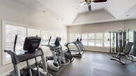 Located at 101 Shore Lake Dr in Greensboro, NC, this fitness center is a well-appointed, single-floor facility designed to support a full range of workout routines. The space features vaulted ceilings with ceiling fans, creating an open and airy atmosphere throughout. Large windows fitted with plantation shutters line the walls, allowing ample natural light to fill the room while offering views of the surrounding greenery.

The cardio zone is equipped with multiple True-brand treadmills, elliptical trainers, and a recumbent stationary bike, all arranged to take advantage of the natural light from the windows. A floor-to-ceiling mirror wall runs along one side of the space, visually expanding the room and supporting proper form during workouts. A wall-mounted flat-screen television adds convenience for entertainment during exercise sessions.

The strength training area includes a multi-station cable machine, a dumbbell rack with a wide range of weights, and a weight bench. A bold navy blue accent wall anchors the functional training zone, which is outfitted with a captain's chair, stability balls in multiple sizes, a balance board, resistance tools, and a dedicated storage rack. Rubber flooring covers the entire space, ensuring safety and durability. A restroom is conveniently accessible directly from the fitness floor.