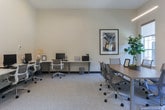 Located at 1800 Deerfield Pt in Milton, Georgia, this single-floor professional office space offers a clean, modern design tailored for productivity and collaboration. The interior is finished with neutral white walls, recessed lighting, and light speckled carpeting throughout, creating a bright and focused work environment.

The main workspace features a perimeter workstation layout with multiple computer stations, ergonomic mesh chairs, and generous desk surface area, accommodating a sizeable team. A central conference table provides additional workspace for group discussions or collaborative tasks, complemented by modern seating.

A dedicated meeting or huddle area is defined by a wall-mounted flat-screen television, windows that invite natural light and views of the surrounding greenery, and a small lounge nook furnished with a pair of bold yellow accent chairs and side tables — adding a pop of color and a welcoming touch to the otherwise neutral palette. Framed artwork is displayed thoughtfully throughout the space, enhancing the professional yet approachable atmosphere.

Large glass partition doors connect distinct zones within the office, supporting an open yet organized flow between areas. The overall design balances functionality with aesthetic appeal, making this Milton office space well-suited for a dynamic, modern team.