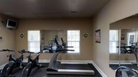Located at 100 Delaney Dr in Jacksonville, NC, this single-floor fitness center offers a dedicated space for residents or members to maintain an active lifestyle. The room is thoughtfully designed to maximize functionality, featuring a selection of cardio equipment including a treadmill and stationary bike, making it well-suited for a variety of workout routines. A large wall mirror spans a significant portion of the room, visually expanding the space and allowing users to monitor their form during exercise. Natural light filters in through multiple windows fitted with blinds, creating a bright and welcoming atmosphere. The dark, rubberized flooring provides both durability and comfort underfoot, ideal for high-impact activity. Motivational wall art adds an energizing touch to the environment. The neutral-toned walls and clean white baseboards contribute to a tidy, well-maintained appearance. Ceiling ventilation ensures proper air circulation throughout the space. Overall, the fitness center presents a practical and comfortable environment designed to support an active and healthy lifestyle.