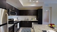 This single-floor space showcases a well-appointed kitchen as its centerpiece, designed with a cohesive modern aesthetic. Rich, dark espresso-toned cabinetry lines the walls from base to upper levels, providing generous storage capacity while creating a bold visual contrast against the light-toned walls and floors. The countertops feature speckled granite surfaces, adding texture and elegance to the workspace.

The kitchen is equipped with a full suite of stainless steel appliances, including an electric range, built-in microwave, and dishwasher, all seamlessly integrated into the cabinetry layout. A stainless steel sink is positioned beneath upper cabinets, keeping the workflow functional and organized.

A defining architectural feature is the tray ceiling above the kitchen area, which adds dimension and visual interest to the space. Recessed track lighting is installed within this elevated ceiling detail, providing focused, adjustable illumination throughout the kitchen. Light-toned tile flooring runs through the area, complementing the overall palette. The kitchen opens toward an adjacent area, suggesting a connected, open-concept layout that enhances the sense of space and flow throughout the unit.