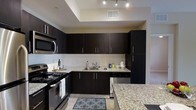 This single-floor space showcases a well-appointed kitchen as its centerpiece, designed with a cohesive modern aesthetic. Rich, dark espresso-toned cabinetry lines the walls from base to upper levels, providing generous storage capacity while creating a bold visual contrast against the light-toned walls and floors. The countertops feature speckled granite surfaces, adding texture and elegance to the workspace.

The kitchen is equipped with a full suite of stainless steel appliances, including an electric range, built-in microwave, and dishwasher, all seamlessly integrated into the cabinetry layout. A stainless steel sink is positioned beneath upper cabinets, keeping the workflow functional and organized.

A defining architectural feature is the tray ceiling above the kitchen area, which adds dimension and visual interest to the space. Recessed track lighting is installed within this elevated ceiling detail, providing focused, adjustable illumination throughout the kitchen. Light-toned tile flooring runs through the area, complementing the overall palette. The kitchen opens toward an adjacent area, suggesting a connected, open-concept layout that enhances the sense of space and flow throughout the unit.