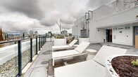 This rooftop terrace, located at 825 Fort St in Victoria, BC, offers a stunning outdoor retreat with panoramic views of the cityscape. The space is thoughtfully designed to maximize comfort and functionality, featuring a variety of seating options including Adirondack chairs, loungers, and communal tables. Large planters with young trees and vegetation add a touch of nature to the urban environment.

The terrace is equipped with several amenities, including a swimming pool, raised garden beds for community gardening, and designated areas for relaxation and socializing. Safety is ensured by glass railings that preserve the expansive views while providing protection. The flooring consists of durable concrete tiles, creating a clean and modern aesthetic.

A notable feature is the mix of materials used, such as white-painted structures, natural wood elements, and metal accents, which contribute to a contemporary yet inviting atmosphere. The layout is spacious and open, allowing for easy movement and flexible use of the area. Despite the cloudy weather depicted, the terrace's design suggests it would be an ideal spot for both sunny day activities and evening gatherings, offering residents a versatile outdoor living space high above the city streets.