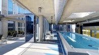 This striking rooftop pool facility offers a seamless blend of indoor and outdoor environments through an expansive glass wall system fitted with large folding panels that open onto an exterior terrace. The lap pool runs along one side of the space, finished in a blue and white mosaic tile pattern that reflects the abundant natural light flooding in through the floor-to-ceiling glazing. Cylindrical concrete columns punctuate the poolside deck at regular intervals, supporting a raw concrete ceiling through which large metallic ductwork runs prominently, contributing to the space's industrial-modern aesthetic. The pool deck is finished in light-toned tile, with seating areas positioned along the length of the pool to allow for relaxation while taking in elevated city views. Beyond the glass panels, an outdoor terrace provides additional lounging space complete with deck chairs. The surrounding urban skyline, visible on multiple sides, reinforces the elevated position of this facility and creates a dramatic backdrop. The overall ambiance is clean, contemporary, and light-filled, combining utilitarian architectural elements with an open, airy atmosphere.
