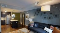 This single-floor space showcases a thoughtfully designed interior that blends contemporary aesthetics with a welcoming ambiance. The living area serves as a central focal point, anchored by a deep navy sofa adorned with eclectic accent pillows and set against a striking slate-blue accent wall. Decorative wire botanical sculptures add an artistic, three-dimensional element to the wall, enhancing the room's visual depth and modern character.

Dark hardwood flooring runs throughout, providing a rich, warm contrast to the cooler tones of the walls and furnishings. A large drum pendant light and a table lamp with a metallic base provide layered lighting options, creating a versatile atmosphere suitable for both daytime and evening use.

An area rug anchors the seating arrangement, while a small side table adds functional elegance near the sofa. Adjacent to the living space, a partial view into what appears to be a kitchen or bar area reveals bar-height seating, pendant lighting, and an olive-toned accent wall, suggesting a seamlessly connected open-concept layout. A wood-toned interior door provides access to additional areas of the space, contributing to the overall cohesive and well-curated design.