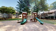 This community space includes an outdoor playground and an adjoining interior living area. The playground is anchored by a substantial wooden play structure featuring two elevated platforms connected by a walkway, two green slides, and small roofed towers. The structure sits within a gravel safety surface, bordered by green lawn and framed by mature trees, including tall evergreens, that provide natural shade. Residential buildings are visible in the surrounding area, situating the playground within a close-knit community setting.

The interior living space transitions from the outdoors through a sliding glass door fitted with vertical blinds, allowing natural light to filter in and providing direct access to an outdoor balcony. The living room features neutral-toned carpet, light gray walls, and a textured ceiling. A fireplace with a dark surround serves as a focal point, complemented by colorful abstract artwork. The layout flows through a hallway connecting to additional rooms, with a closet and utility storage accessible nearby. The entry foyer features dark hardwood flooring, offering a clean contrast to the carpeted living areas. The overall ambiance is understated and functional.