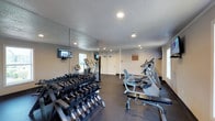 This fitness center offers a comprehensive workout environment designed to accommodate a wide range of exercise routines. The space is laid out on a single floor with durable, dark rubber flooring throughout, providing traction and cushioning ideal for high-activity use. Recessed lighting illuminates the room evenly, complemented by multiple windows that allow natural light to fill the space and provide ventilation.

A large mirrored wall section spans a significant portion of the room, visually expanding the space and allowing users to monitor their form during workouts. The cardio zone is well-stocked, featuring treadmills, elliptical trainers, and a stationary bike, several of which are equipped with individual digital displays. Multiple wall-mounted flat-screen televisions are positioned throughout the room for entertainment during exercise sessions.

The free weights area includes a multi-tier dumbbell rack with a wide range of weight options, paired with a workout bench for strength training. A dedicated functional training station houses a TAGfit rack stocked with medicine balls ranging from 8 to 20 pounds, along with stability balls for core and balance work. Two access doors are visible along one wall, and a posted fitness center rules sign indicates this is a shared community amenity. The textured ceiling and crown molding add a finished, polished look to the overall space.
