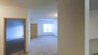 This single-floor space presents a straightforward and functional layout characterized by clean, neutral design throughout. The living area is finished with wall-to-wall carpet in a light, neutral tone, providing a soft underfoot feel and a cohesive look across the open floor plan. The walls are painted in a crisp off-white, contributing to a bright and airy atmosphere throughout the interior.

Natural light enters through a window positioned along the far wall of the main living area, illuminating the space and enhancing its open feel. The ceilings are standard height and smoothly finished, maintaining a clean and uncluttered overhead plane.

A light wood-toned interior door is visible within the space, consistent with the overall warm, neutral palette. An additional doorway is visible to one side, suggesting connectivity to adjacent rooms or areas within the unit. The architectural layout appears straightforward, with partitioning walls that define separate zones while maintaining a sense of openness.

Overall, the space offers a clean, move-in-ready environment with a simple, neutral aesthetic that provides a versatile backdrop suitable for a variety of furnishing and decorating styles.