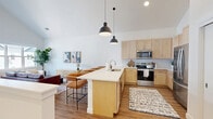 Located at 239 Water Street in West Bend, WI, this thoughtfully designed one-bedroom, one-bathroom residence offers a bright and modern living experience across a single floor. The open-concept kitchen and living area serves as the heart of the home, highlighted by dramatic vaulted ceilings that create an airy, expansive atmosphere. The kitchen is well-appointed with light maple cabinetry, white quartz countertops, a stainless steel appliance suite including a refrigerator, range, and microwave, a white tile backsplash, and a central island with pendant lighting and bar seating — ideal for both cooking and casual dining. The adjoining living area is bathed in natural light from large windows overlooking the neighborhood, and features warm hardwood-style flooring that carries throughout the entire home. The bedroom is a comfortable, well-proportioned retreat with a window providing natural light and outdoor views. The full bathroom includes a light wood vanity with a white countertop, a large mirror, and clean, minimalist finishes. A dedicated closet and hallway complete the layout. The overall design aesthetic is clean and contemporary, blending warm wood tones with crisp white walls for a welcoming and functional living space.