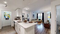 This single-floor residence is designed with a clean, contemporary aesthetic, centered around an open-concept layout that seamlessly connects the kitchen, dining, and living areas. The kitchen is a standout feature, equipped with stainless steel appliances, white cabinetry with sleek hardware, and an expansive quartz-topped island with an undermount sink — providing both functional workspace and an informal gathering point. Track lighting overhead enhances visibility, while wood-style plank flooring runs consistently throughout the home, lending warmth and visual continuity.

The living area is generously proportioned, receiving abundant natural light through large windows dressed with floor-length curtains. A glass-paneled entry door further brightens the space. The two bedrooms are well-sized, each featuring ceiling fans, windows, and easy access to storage. One bedroom connects directly to a spacious walk-in closet outfitted with wire shelving systems, offering practical organization capacity.

The full bathroom features a white quartz vanity, a large wall mirror, and wood-style flooring that complements the rest of the home. A dedicated in-unit laundry area with a full-size washer and dryer adds everyday convenience, completing this well-appointed residence.