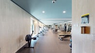 This contemporary fitness center offers a thoughtfully designed, elongated layout that provides ample space for a full range of workout activities. The facility features a sleek teal-painted ceiling accented by long linear LED lighting strips, lending the space a modern and energizing ambiance. Durable rubber flooring spans the entire area, providing both safety and comfort underfoot.

The cardio zone is positioned along a windowed wall, where treadmills and elliptical trainers face large windows with roller shades, allowing natural light to filter in and offering views of the outdoors. This area transitions seamlessly into a strength training section, which houses a comprehensive selection of cable machines and multi-use weight stations arranged for efficient use of the floor space.

A dedicated free weights area features a full rack of dumbbells neatly organized by weight, a flat-adjustable bench, medicine balls, and a rowing machine. A wall-mounted flat-screen television serves this zone, making it ideal for functional training. Full-length mirrors line one wall, visually expanding the space and assisting users with form.

The warm wood-paneled accent walls add a natural, refined element that balances the industrial equipment aesthetic. The overall design is clean, modern, and purposeful, making this fitness center a comprehensive and inviting space for all types of training.