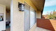 This single-floor residential unit offers a practical and comfortable layout with several well-defined living areas. The exterior features a textured stucco facade with a classic wall-mounted lantern light fixture and large sliding windows that bring in natural light.

Inside, the kitchen is efficiently designed with a galley-style layout, featuring white cabinetry, dark granite-style countertops, and a bright overhead lighting panel. White appliances and ample cabinet storage make this a highly functional cooking space.

The living area is generously sized, finished with neutral carpet and warm beige walls beneath a textured ceiling. Large vertical blinds frame a sliding glass door that opens directly onto a private balcony, which features a wooden railing and pleasant views of surrounding greenery and neighboring rooftops.

An additional room, accessible through a doorway from the main living area, features hardwood-style flooring and a large sliding glass door with vertical blinds, offering further access to natural light and outdoor views.

Throughout the unit, the design is clean and understated, with neutral tones and practical finishes that create a calm, livable atmosphere. The combination of indoor-outdoor access via multiple sliding glass doors is a consistent and notable feature of this space.