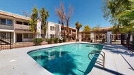 Located at 1022 E Narramore Ave in Buckeye, Arizona, this outdoor community pool area offers a welcoming and functional amenity space designed for relaxation and recreation under the Arizona sun. The centerpiece is a generously sized swimming pool with clear turquoise water, finished with smooth concrete pool decking that provides ample space for lounging and foot traffic. Stainless steel entry rails offer easy pool access, and a life preserver ring is conveniently positioned poolside for safety.

A covered pergola structure with thick stucco columns anchors one corner of the pool deck, providing a shaded retreat for seating and gathering. The surrounding landscape is richly planted with mature palm trees, tall weeping trees, and lush shrubs that add both privacy and a verdant, resort-like atmosphere. The entire pool area is enclosed by a sturdy wrought iron security fence, ensuring a safe and defined perimeter.

The adjacent multi-story building features classic Southwestern stucco architecture with a warm terracotta tile roofline, exterior staircases, and balconies that overlook the pool courtyard. Decorative tile accents and architectural arched details reinforce the regional aesthetic. Under the broad open skies of Buckeye, this outdoor space balances practicality with a pleasant, community-oriented ambiance.