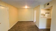 This single-floor space offers a clean, functional layout designed for comfortable everyday living. The main living area is an open, well-proportioned room featuring durable wood-look plank flooring that runs throughout, adding warmth and a modern touch to the interior. The walls are painted in a soft, neutral yellow tone, creating a bright and welcoming atmosphere. A textured popcorn ceiling with a centrally mounted light fixture provides overhead illumination, keeping the space well-lit. White baseboards frame the room neatly, complementing the overall light color palette. A doorway from the living area leads to an additional room, suggesting a connected layout that supports flexible use of space. Adjacent to the living area, the kitchen is partially visible and features white cabinetry, a white range, and a light-colored countertop, offering a practical cooking space with a clean, classic aesthetic. The layout's open sightlines between the living and kitchen areas contribute to a sense of connectivity and ease of movement throughout the unit. Overall, this space presents a straightforward, move-in-ready interior with consistent finishes and a practical design suited for a variety of living arrangements.