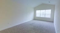 This single-floor space is defined by its striking vaulted ceiling, which rises dramatically to create an impressive sense of openness and volume. The architectural detail of the angled ceiling adds character and visual interest, making the room feel notably spacious and airy. Neutral-toned wall-to-wall carpeting covers the floor throughout, providing a soft, comfortable surface underfoot while complementing the clean, bright aesthetic of the space. The walls are finished in a crisp, bright white, contributing to an overall light and fresh atmosphere. A large multi-pane window fitted with horizontal blinds is positioned prominently along one wall, allowing generous amounts of natural light to fill the interior. The window also provides a view to the exterior, enhancing the connection between indoors and outside. The combination of the vaulted ceiling, abundant natural light, and neutral finishes creates a welcoming and versatile environment that can accommodate a variety of furniture arrangements and uses. The clean lines and minimalist design of the space lend it a timeless, adaptable quality suited to a range of occupants and interior styles.