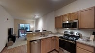 This single-floor space showcases an open-concept layout where the kitchen seamlessly connects to the dining and living areas, creating a cohesive and functional environment. The kitchen is well-appointed with stainless steel appliances, including an electric range, built-in microwave, and dishwasher. Warm-toned maple cabinetry provides ample storage, while the granite countertops in soft earth tones add a touch of elegance and durability to the workspace. A double-basin sink is positioned along the peninsula, which also serves as a natural divider between the kitchen and the adjoining dining area. Track lighting overhead ensures the kitchen is well-lit, complementing the warm ambient tones throughout. Light hardwood-style flooring runs through the kitchen, transitioning into carpeted areas in the dining and living spaces. The dining area accommodates a full table and chairs, and a glass door at the far end floods the space with natural light while offering views of a greenery-filled outdoor area. The overall ambiance is warm, practical, and inviting, suited for everyday living.