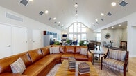 Located on Hancock Expy in Colorado Springs, CO, this single-story community clubhouse offers an expansive, open-concept layout designed for both relaxation and socializing. The centerpiece of the space is a dramatic vaulted ceiling that rises to a striking peak, complemented by a floor-to-ceiling gabled window wall that floods the interior with natural light. Light hardwood flooring flows consistently throughout, lending warmth and cohesion to every area.

The main gathering area features a generous seating arrangement anchored by a large leather sectional sofa, a wooden coffee table, and comfortable accent chairs — all arranged around a welcoming lounge setting. Adjacent to this is a formal dining area with a solid wood table and matching chairs, ideal for group gatherings. The fully equipped kitchen showcases dark espresso cabinetry, granite countertops, a stainless steel refrigerator, and a central island for casual dining or prep work.

Near the entrance, a bright foyer with soaring ceilings and large glass double doors creates an inviting welcome. A cozy lounge nook nearby features a fireplace, wall-mounted TV, and additional seating. A dedicated game room provides a lively retreat, complete with a full-size pool table, pendant lighting, high-top seating, and windows overlooking the surrounding Colorado landscape.