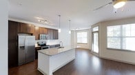 This single-floor residence offers three bedrooms and two bathrooms with a thoughtfully designed open-concept layout. The kitchen serves as a central hub of the home, featuring dark espresso cabinetry that contrasts elegantly with a granite-topped island finished in white. A subway tile backsplash adds a clean, contemporary detail, while stainless steel appliances — including a refrigerator, range, and microwave — provide a fully equipped cooking space. Pendant lighting hangs above the island, complementing the track lighting installed along the kitchen ceiling.

The kitchen flows seamlessly into the living area, which is finished with rich, dark hardwood flooring that extends throughout the main living spaces. The open layout creates a generous sense of space and connectivity between the kitchen, dining, and living zones. Large windows along the far wall allow natural light to fill the area, offering views of the surrounding greenery outside. A glass door provides additional light and access to the exterior. The overall ambiance is modern and welcoming, combining warm wood tones with crisp white walls and contemporary fixtures.