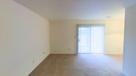This single-floor space offers a clean, functional layout designed for comfortable everyday living. The living area is the centerpiece of the space, featuring wall-to-wall plush carpeting in a neutral tone that lends warmth underfoot. Smooth, off-white walls provide a bright and airy backdrop, creating a versatile canvas for a variety of interior styles. A standout feature of the living area is the full-width sliding glass door dressed with vertical blinds, which serves as both a primary source of natural light and a direct connection to the outdoors. The door allows daylight to pour into the room, enhancing the open and spacious feel of the interior. The ceiling is smooth and standard height, maintaining a clean and uncluttered look throughout. Electrical outlets are conveniently positioned along the walls, offering practical flexibility for furniture arrangement. Overall, the space presents a simple, well-lit, and move-in-ready environment with a calm, neutral ambiance that suits a wide range of uses and personal styles.